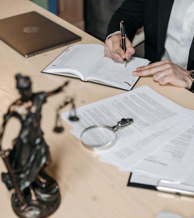 Professional working on documents at desk with legal statue and magnifying glass.